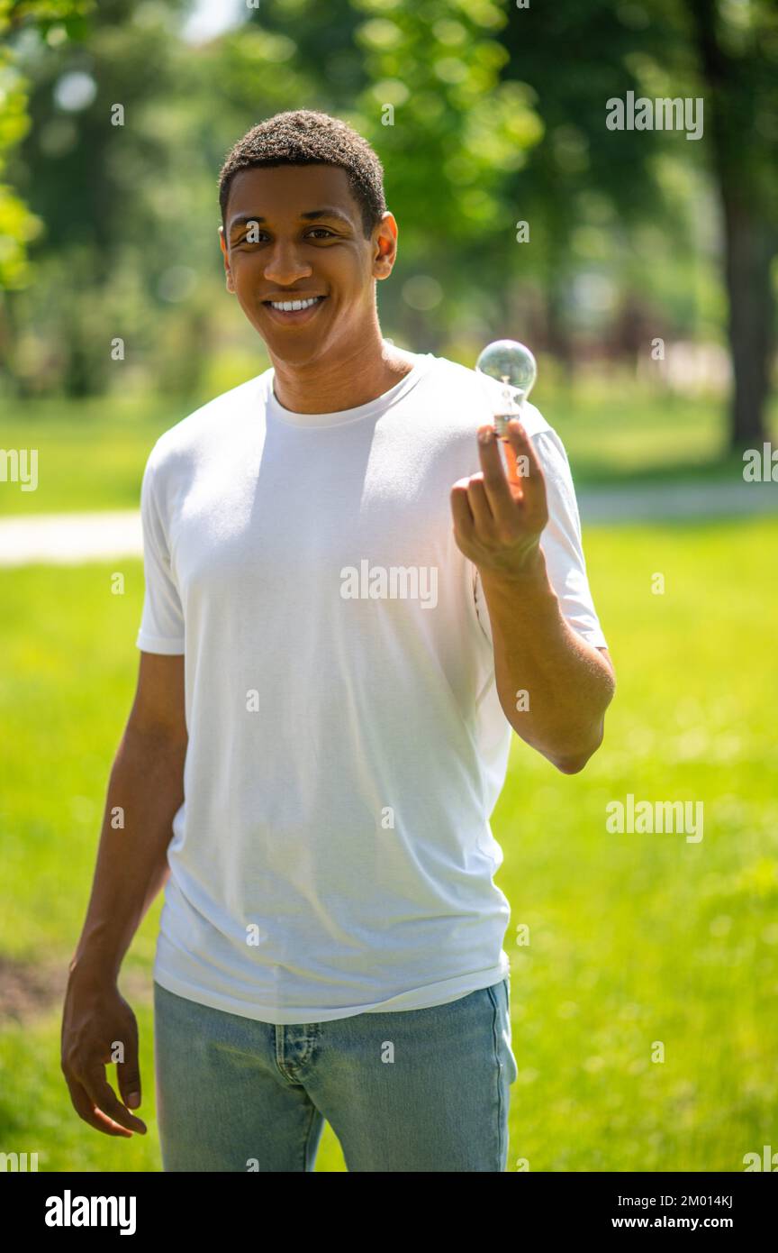 Protection of nature. Dark-skinned guy smiling at camera showing lightbulb standing in park on sunny day. Stock Photo