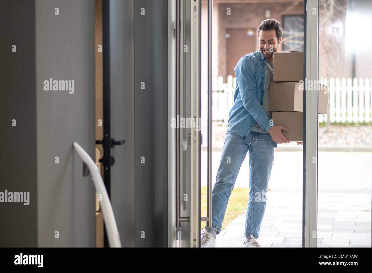 Moving. Young adult smiling man holding stack of boxes looking down propping foot on door