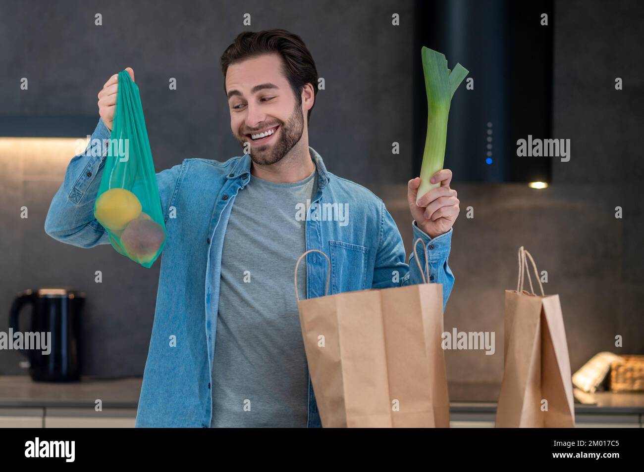 Favorite food. Happy man smiling looking at fruit in raised hand taking out groceries from bags in kitchen. Stock Photo
