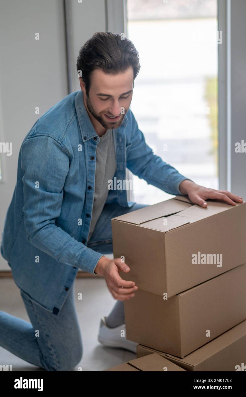 Accuracy. Smiling young adult man kneeling down stacking boxes in hallway of new apartment at daytime. Stock Photo