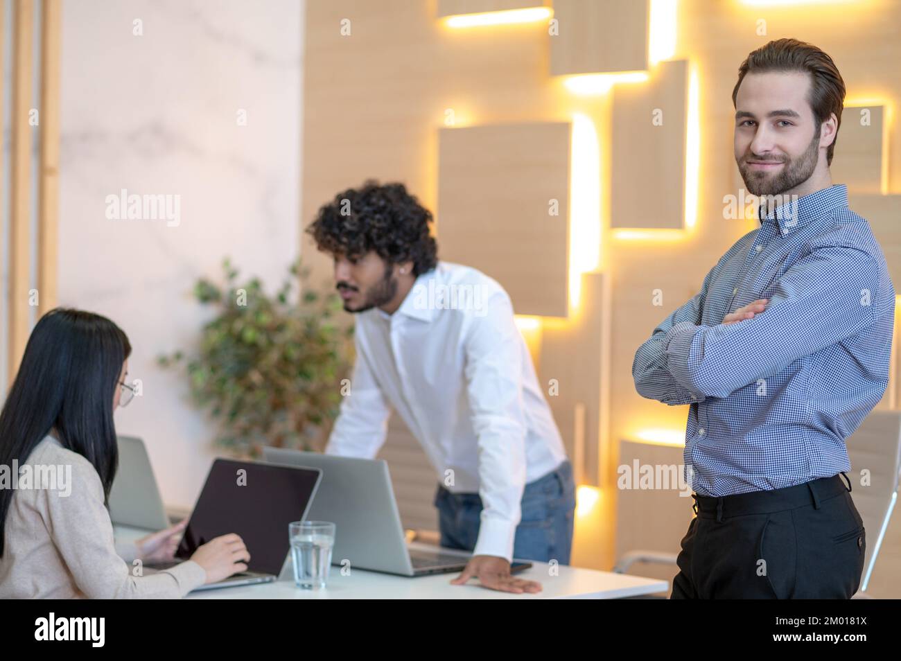 Smiling pleased young office worker standing beside his two busy