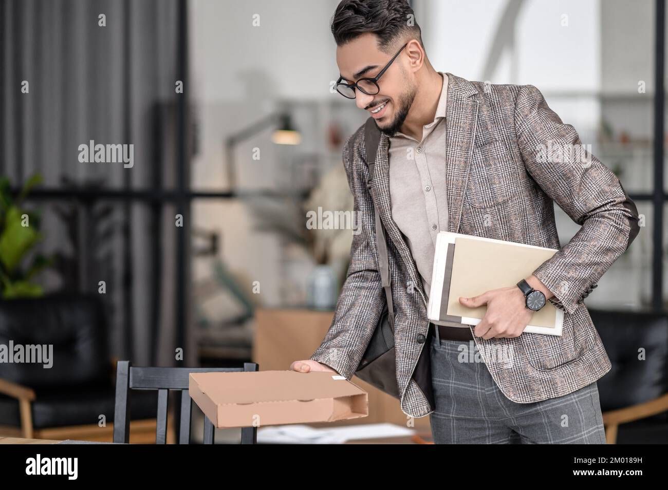 Anticipation. Smiling young bearded man in glasses looking at box in hand holding periodicals standing sideways to camera indoors. Stock Photo