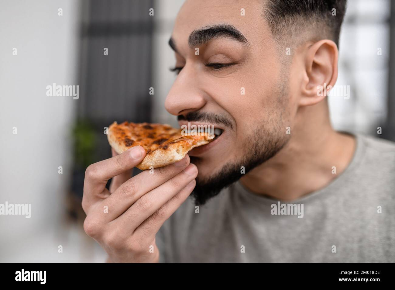 Tasty food. Face of young bearded man sideways to camera biting off delicious fresh pizza indoors. Stock Photo