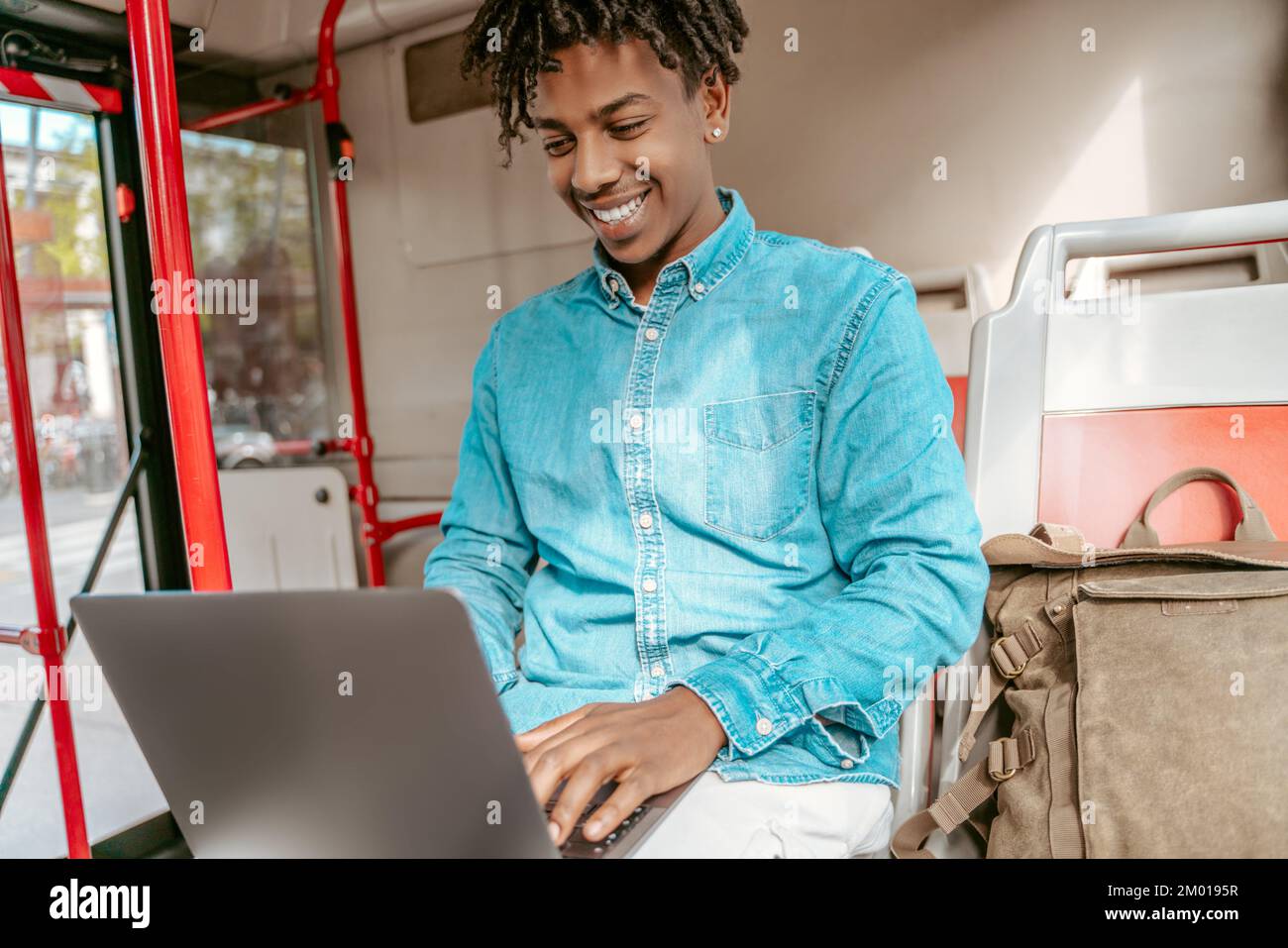 Life Style Cheerful Dark Skinned Guy Typing On Laptop While Sitting Near Backpack In City Bus 