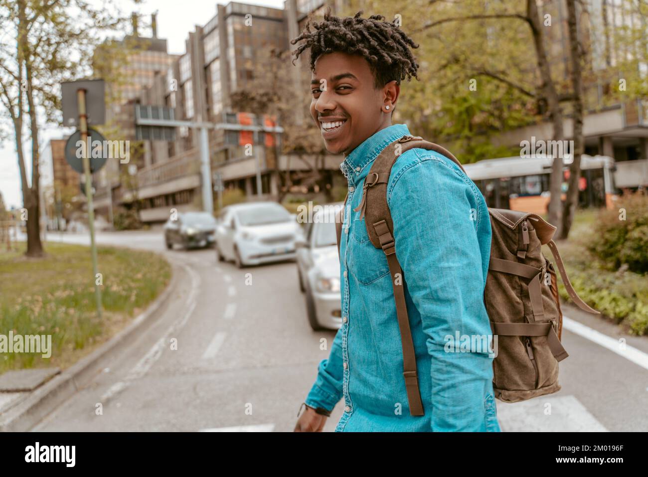 City energy. Dark-skinned stylish guy with backpack smiling confidently at camera crossing road in city. Stock Photo