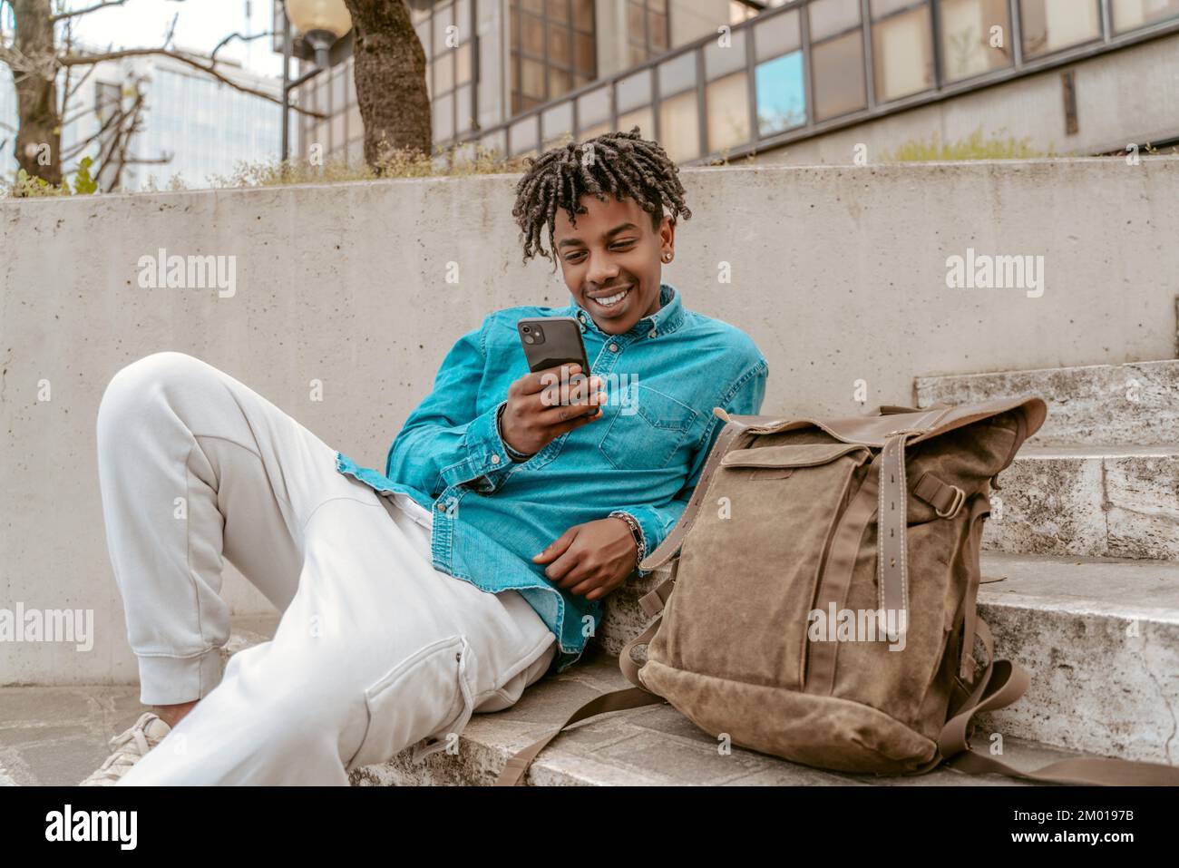 Free time. Dark-skinned joyful guy looking at smartphone sitting relaxed near backpack on steps of city street on fine day. Stock Photo