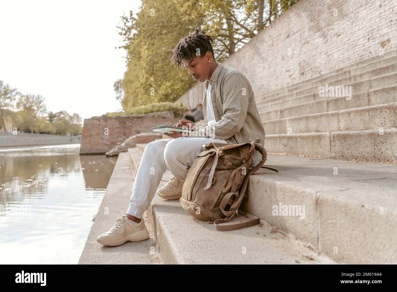 Solitude. Dark-skinned guy looking at tablet touching screen sitting sideways to camera on steps near water. Stock Photo