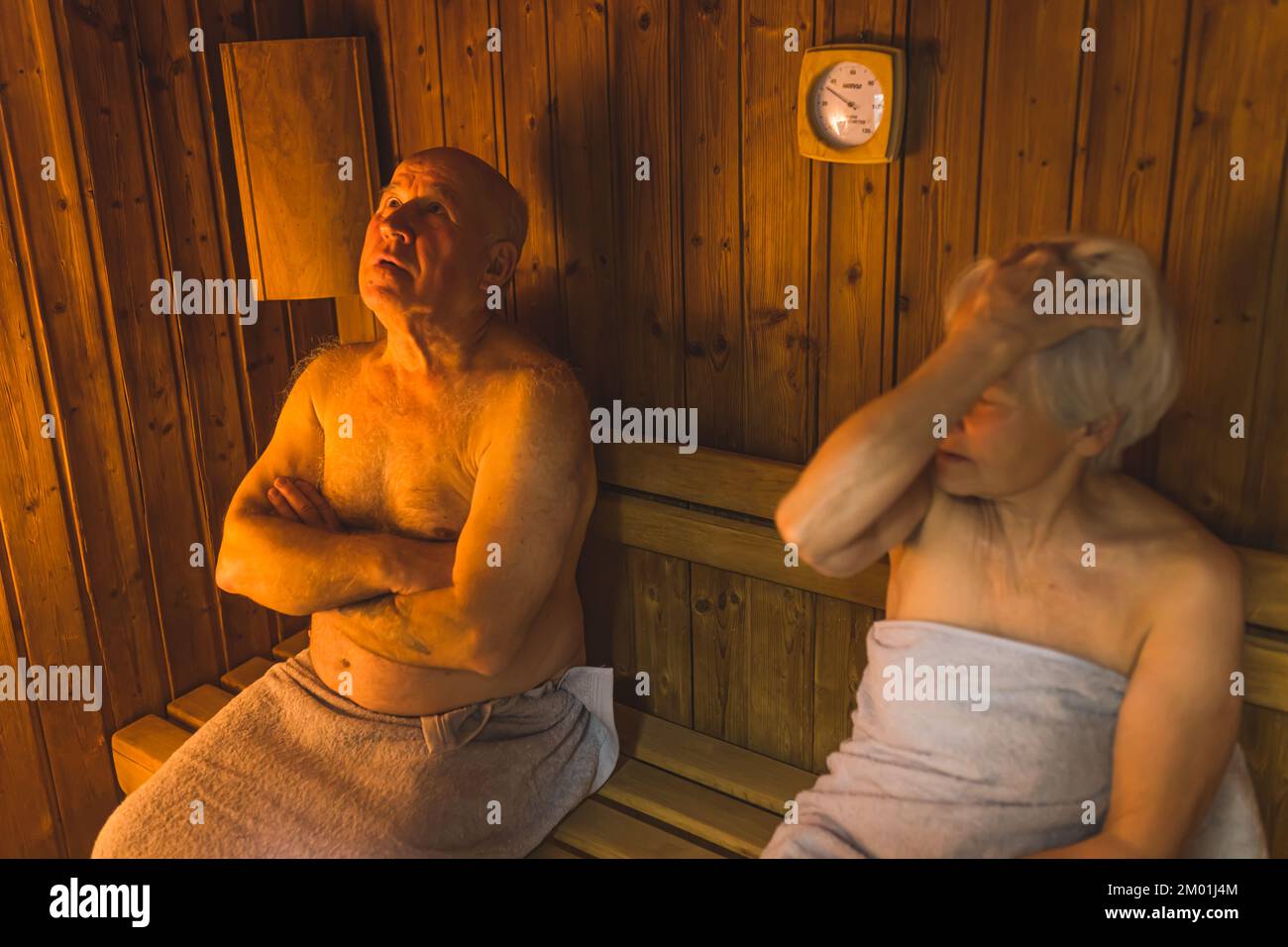 Two caucasian elderly people - bald man and short-haired woman - sit together in a wooden sauna in gray cotton bathrobes. SPA and relaxation concept. High quality photo Stock Photo