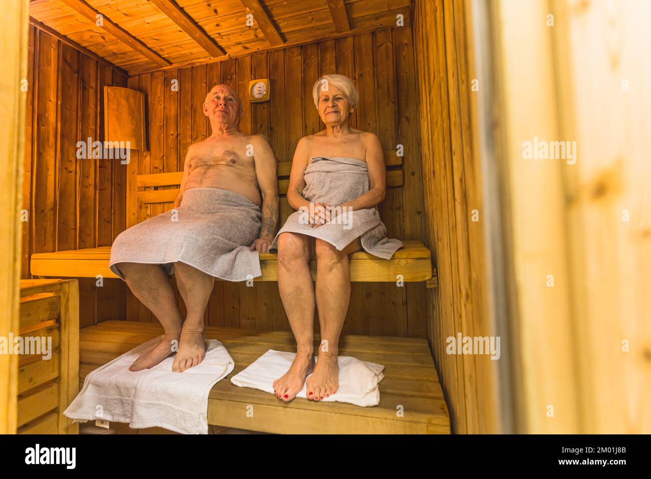 Leisure activity and relaxation concept. Full-length indoor shot of two white grandparents who sit in a wooden sauna with towels under their feet. High quality photo Stock Photo