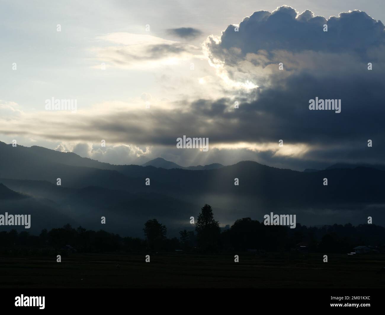 Silhouette of tree and mountain with sunbeam light shoot through the dark cloud to the land at sunrise, Mist covers the forest and mountains at dawn Stock Photo