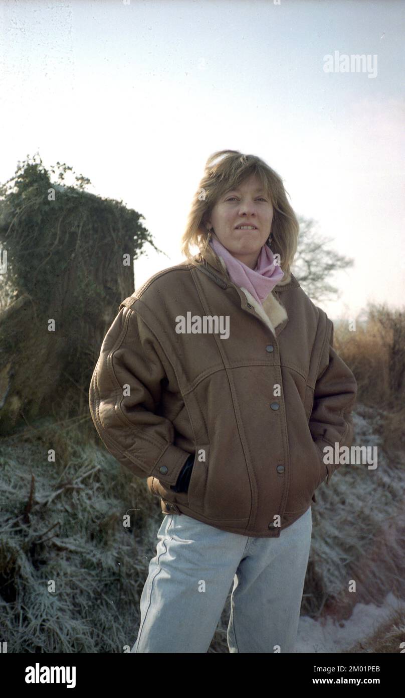 1980s, historical, outside on a frosty morning, with clear skies, a young woman in her mid-twenties standing wearing a brown leather jacket of the era, a bomber jacket with wide shoulders, a pink scarf and faded light blue jeans, England, UK. Bomber or aviator jackets were popular fashion items in the 1980s, particularly after the release of the hit film, Top Gun in 1986. Stock Photo