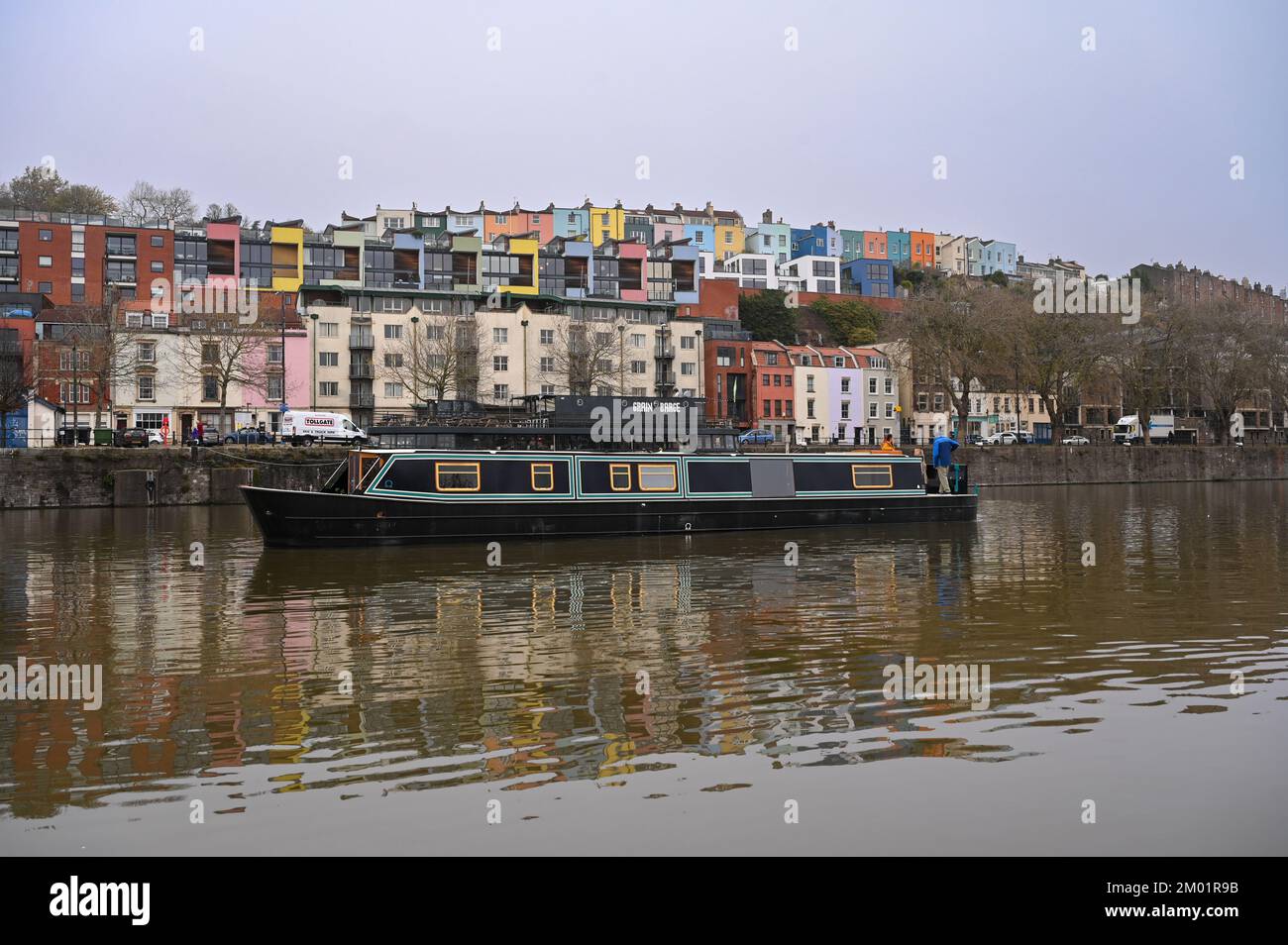 New liveaboard widebeam boat cruising through Bristol docks past a row of colourful houses in the background. Stock Photo