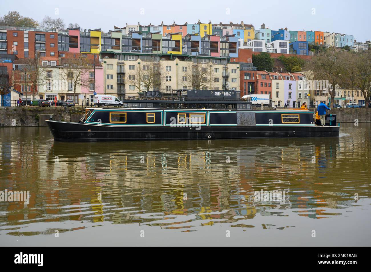 New liveaboard widebeam boat cruising through Bristol docks past a row of colourful houses in the background. Stock Photo