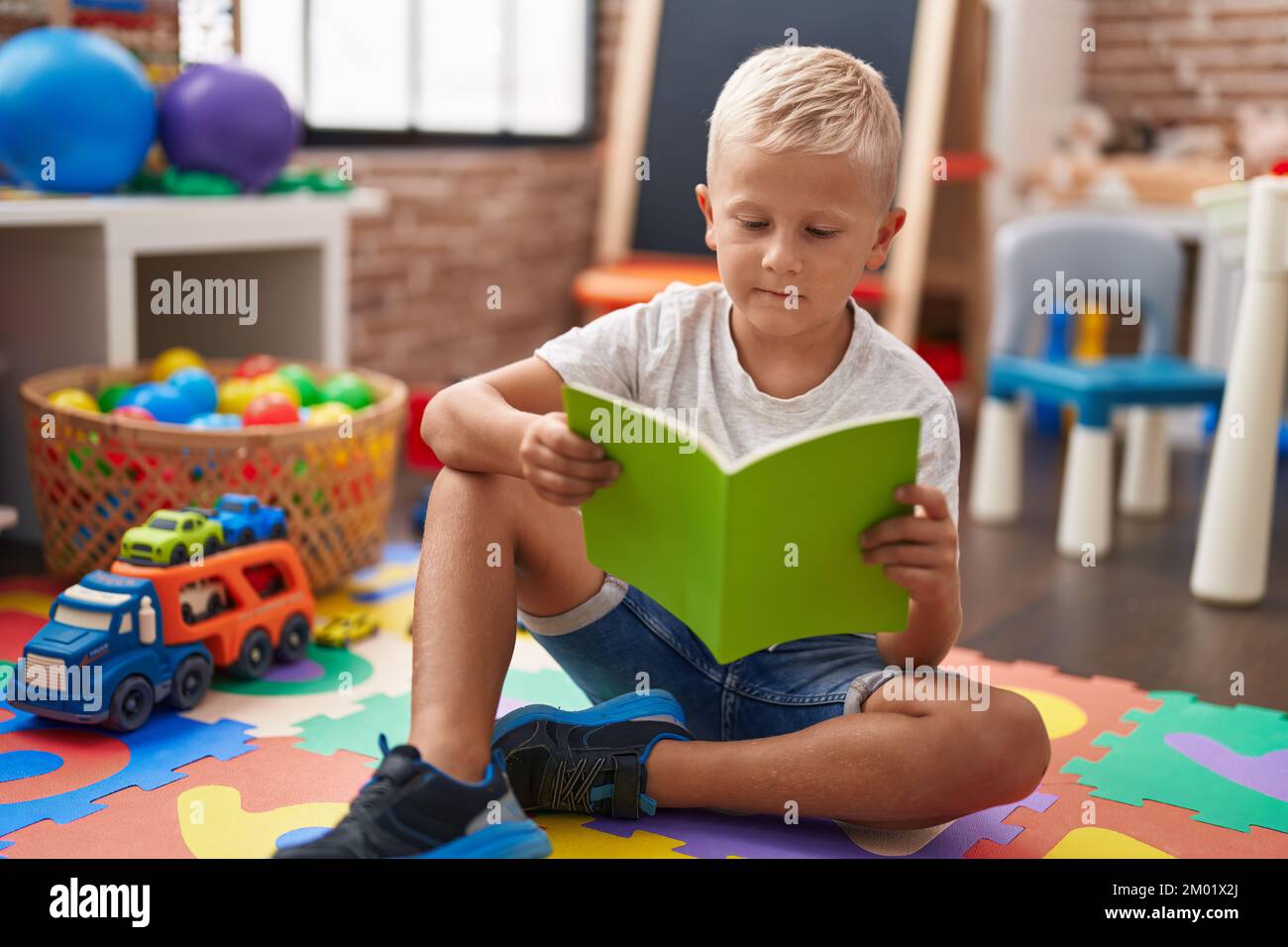 Adorable toddler student reading book sitting on floor at classroom ...