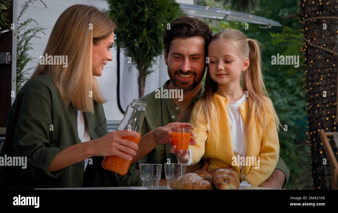 Caucasian family on camping vacation outdoors sitting near van have lunch picnic eat good with croissants mother pouring fresh orange juice into Stock Photo