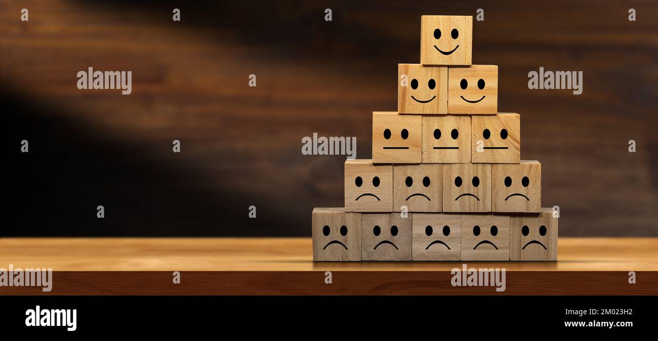 Group of sad, neutral and happy smiley faces on wooden blocks forming a pyramid, above a wooden table or desk with copy space. Stock Photo