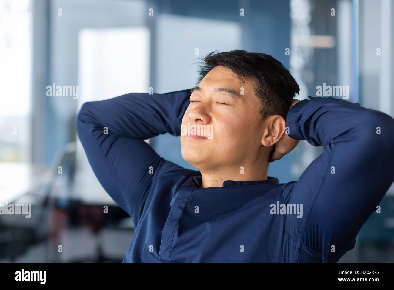 Asian businessman resting in office, man in shirt close-up with hands behind head dreams and visualizes desired plans and achievements. Stock Photo