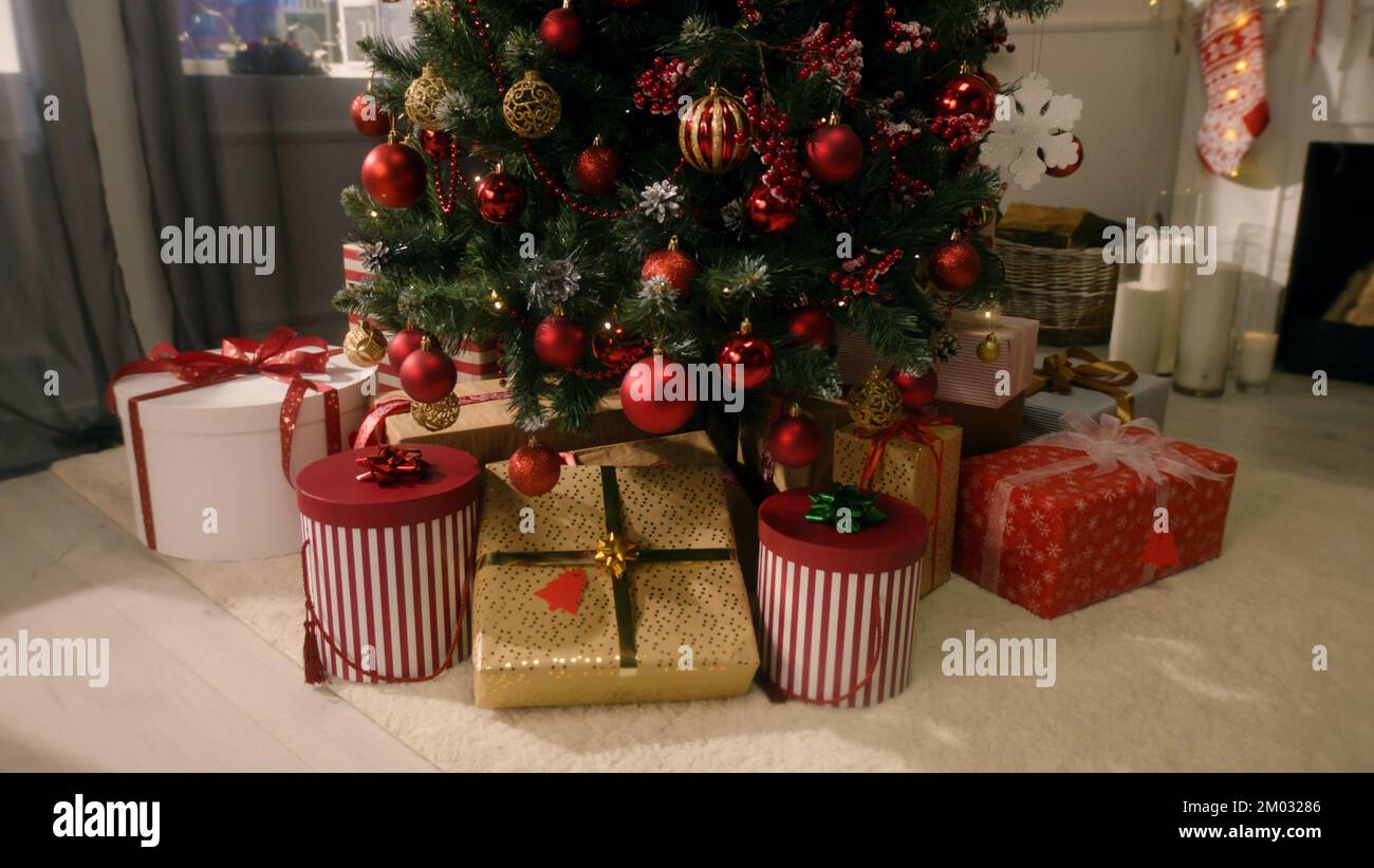 Tree with balls, toys, flashing lights, wrapped gifts and present boxes stands in living room. Christmas and New Year festive interior decoration. Atmosphere in cozy apartment on winter holidays. Stock Photo