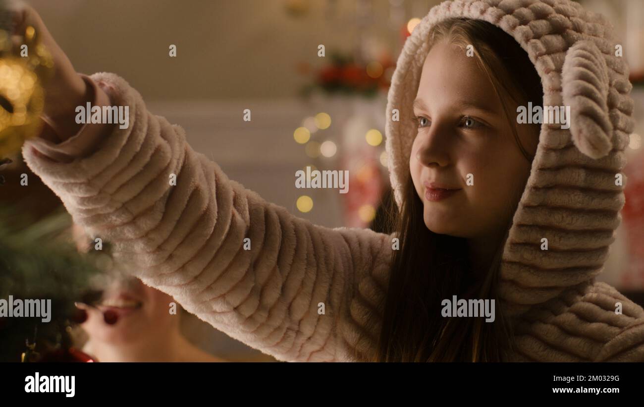 Young girl in pajama choosing balls and decorating Christmas tree with mom. Family preparing living room before Christmas or New Year. Cozy atmosphere of winter holidays. Close-up view. Slow motion. Stock Photo