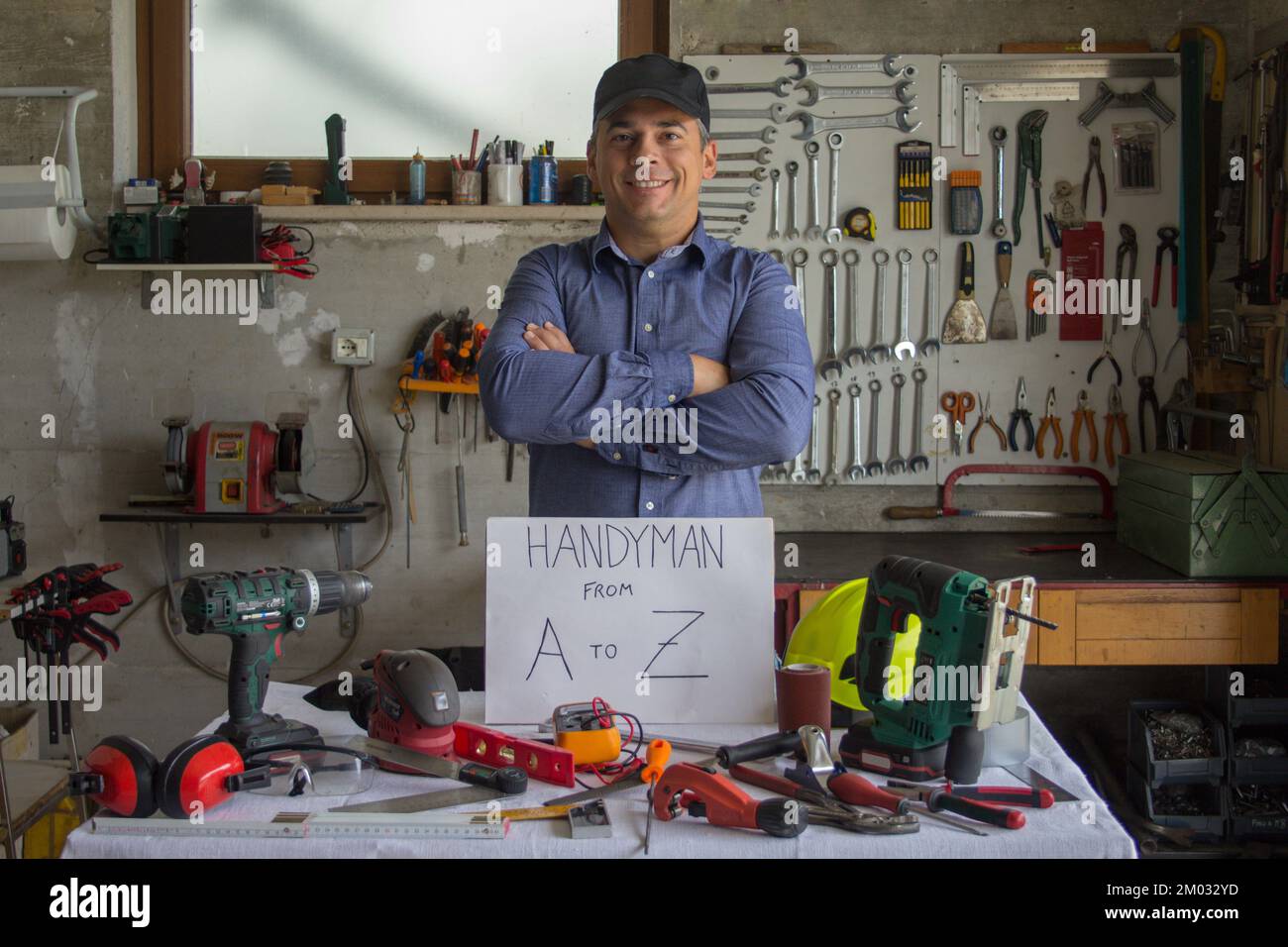 Image of a smiling man in his workshop behind a bench full of work tools with handyman from A to Z written on it. Man who can fix and fix everything Stock Photo