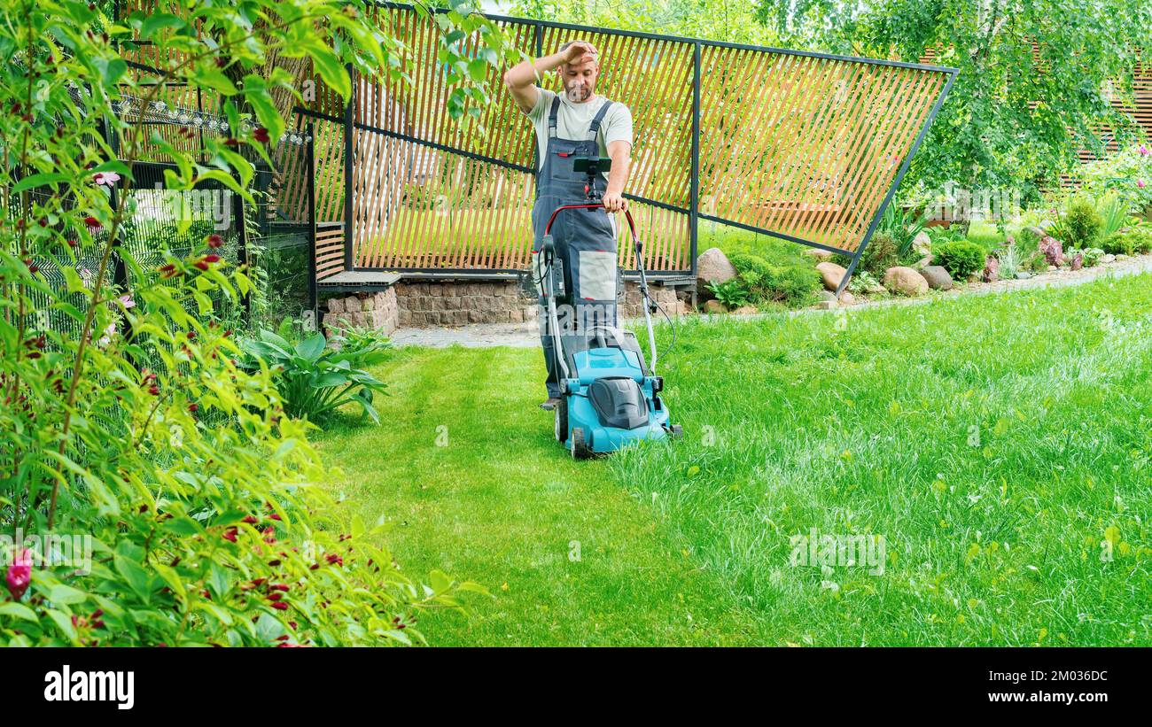 A man mows the lawn with an electric lawn mower in a landscaped garden. A man is tired from the tedious work of mowing the grass on the lawn on a hot Stock Photo