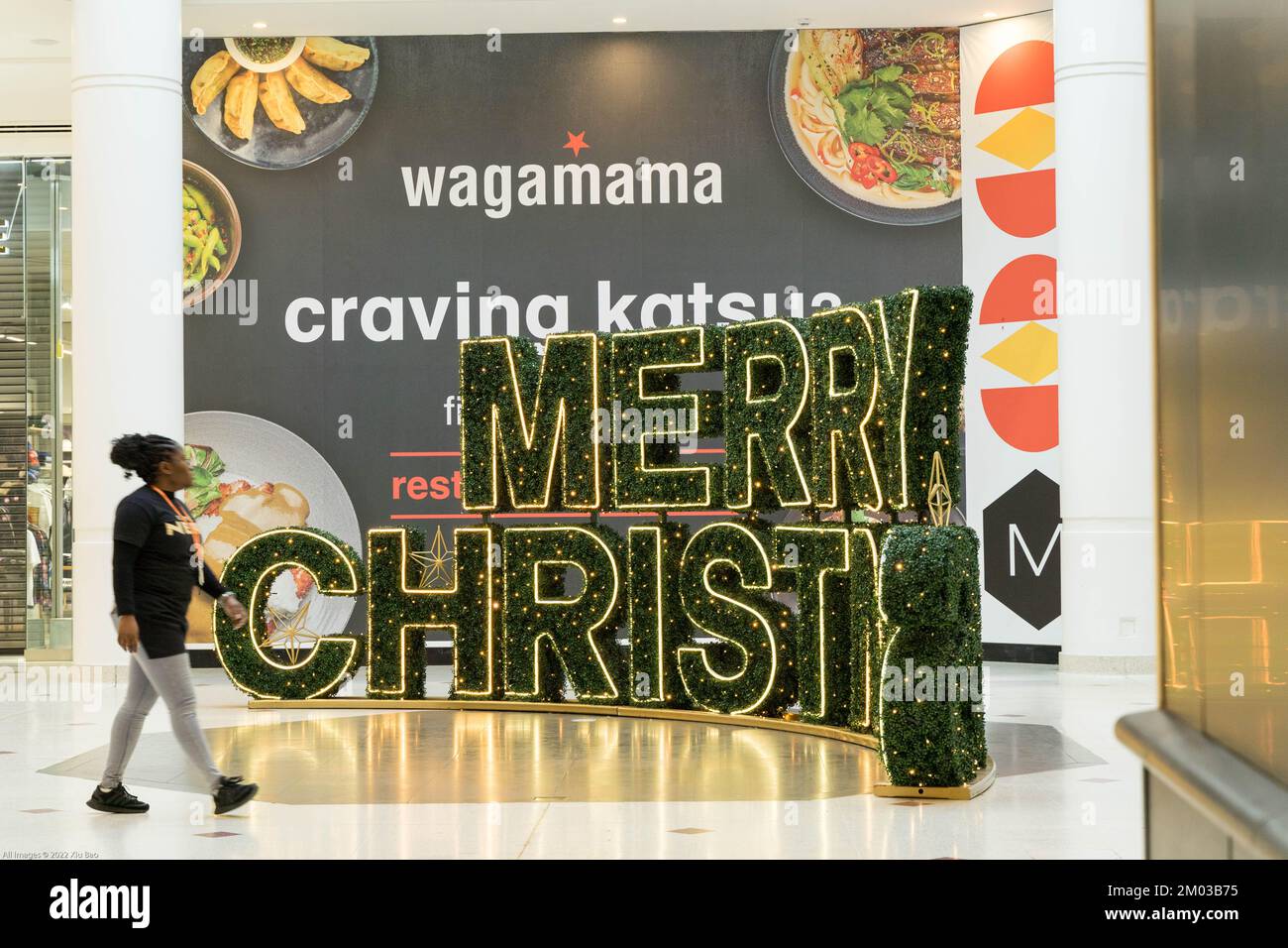 woman walks past a  Merry Christmas tree and lights outside Wagamama restaurant in London shopping mall , England UK Stock Photo