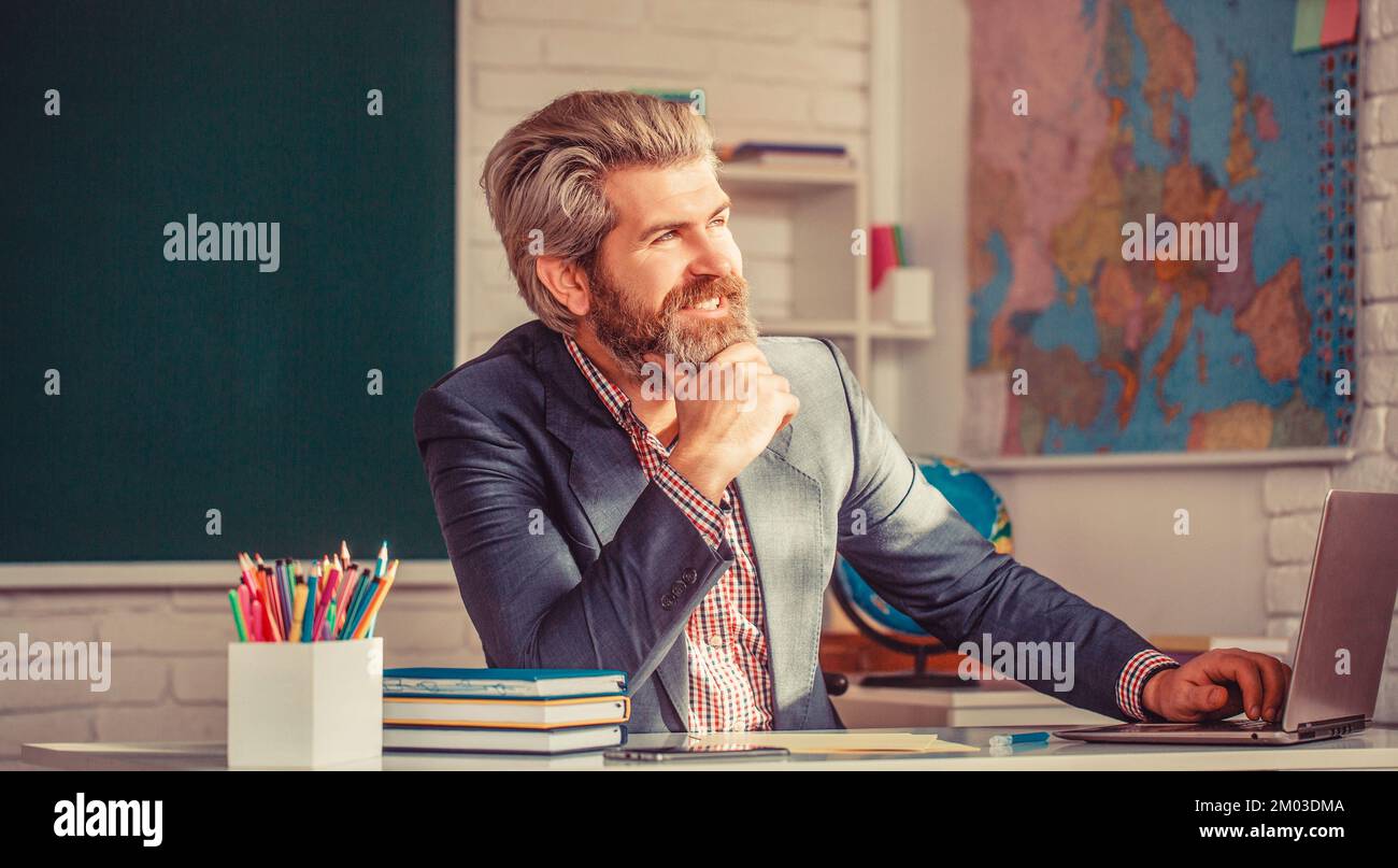 Portrait of confident male teacher in classroom. Young male teacher with laptop in class sits at the table the blackboard in the classroom Stock Photo