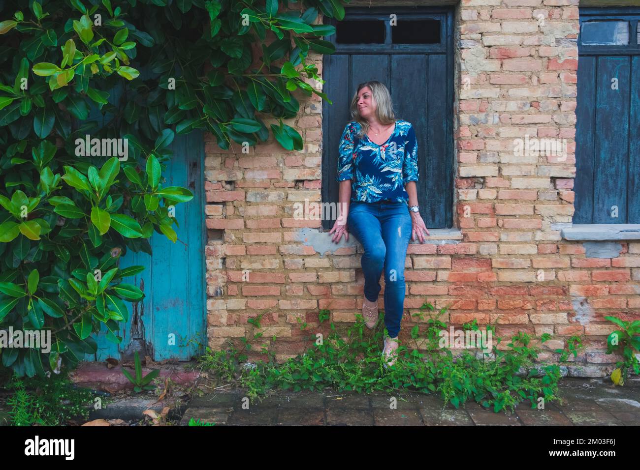 Woman leaning against a window of an old house. Brick facade and leaves around. Aratuipe, Bahia. Stock Photo