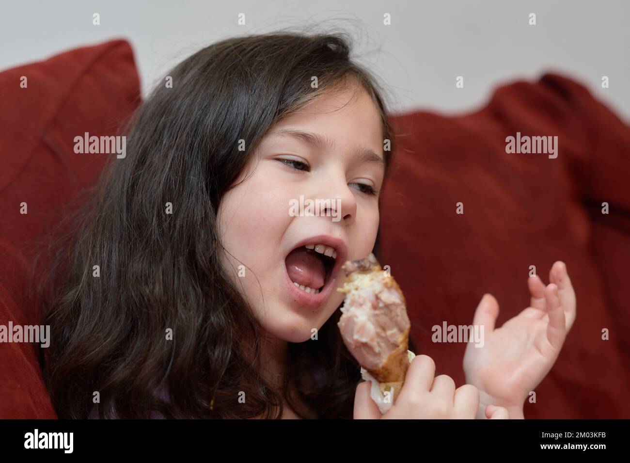 expressive young girl is eating chicken and vegetables for dinner on the couch while watching TV Stock Photo