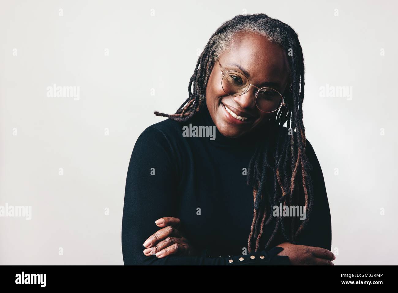 Happy professional woman smiling at the camera while wearing eyeglasses in a studio. Middle-aged woman with dreadlocks standing against a grey backgro Stock Photo
