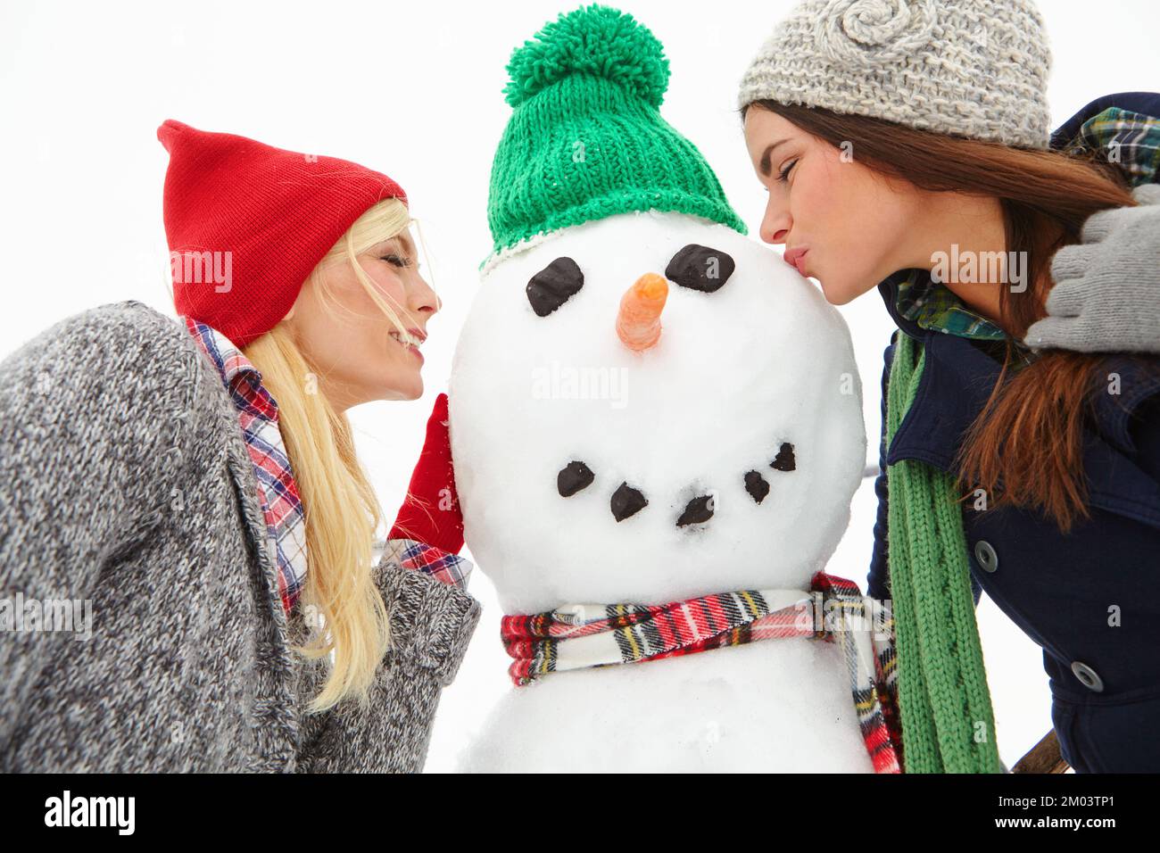 Winter, friends and women kiss snowman enjoying holiday, vacation and festival tradition for Christmas. Nature, happiness and girls having fun Stock Photo