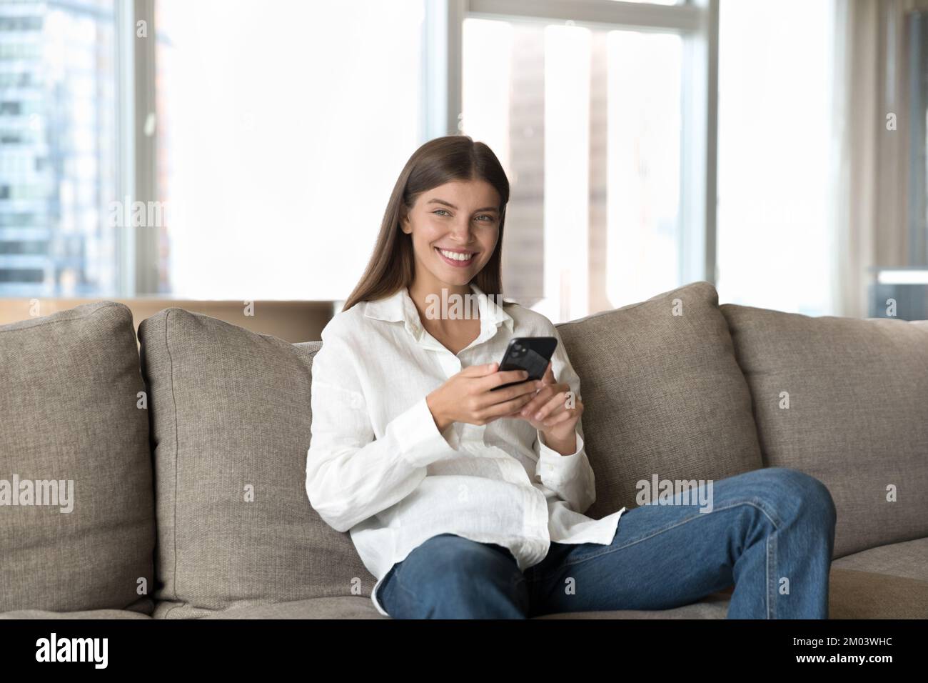Happy positive young cellphone user woman sitting on home sofa Stock Photo