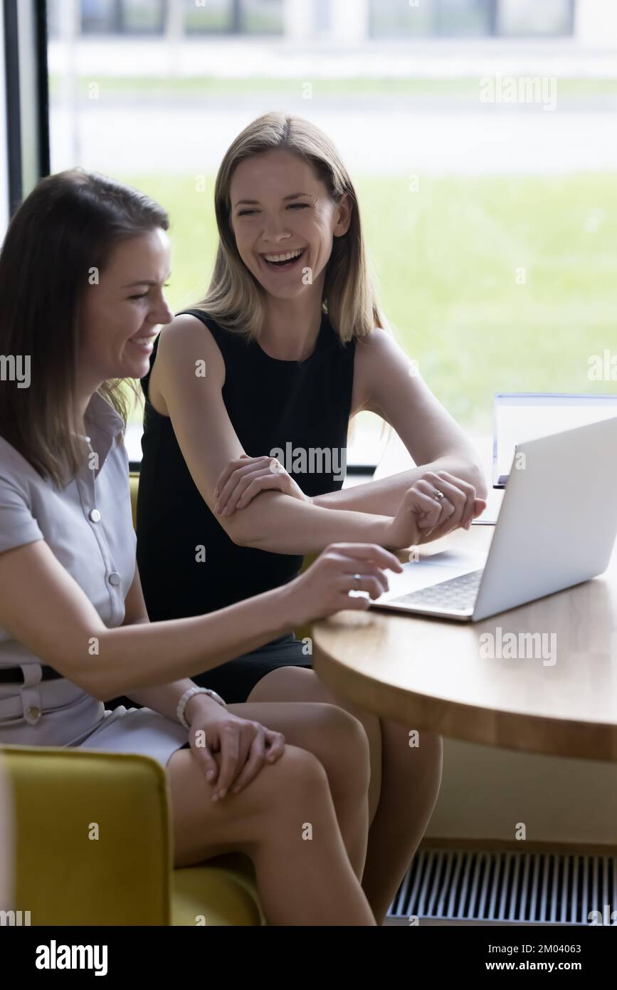 Vertical shot two laughing women sit at table with laptop Stock Photo