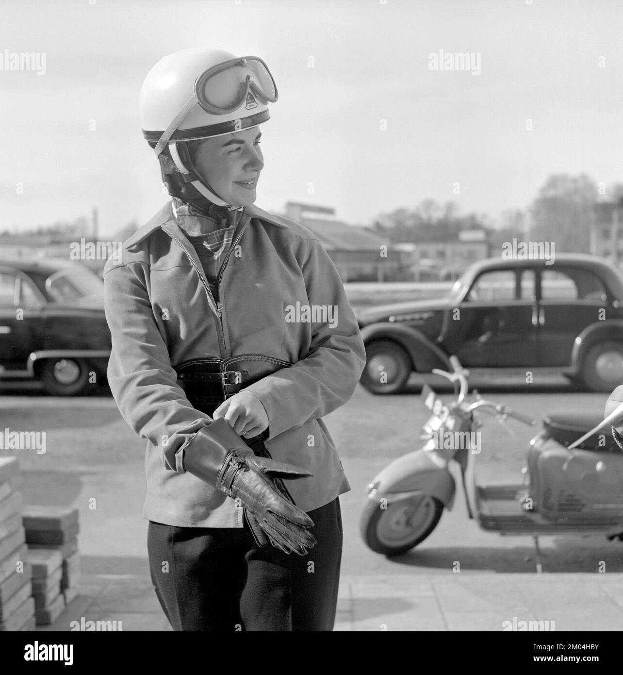In the 1950s. A young woman seen on a german made Puch scooter. The female driver is wearing the typical motorcycle clothing and accessories of the decade, helmet, jacket, kidney belt and gloves. Sweden 1955 Stock Photo