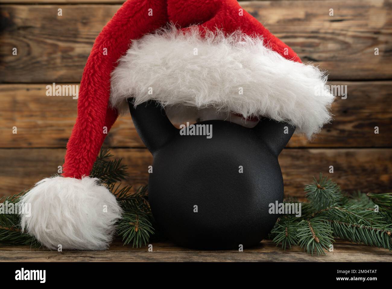 Gym kettlebell in red Santa Claus hat. Exercise equipment as Christmas gift idea. Fitness holiday season, winter workout composition. Stock Photo