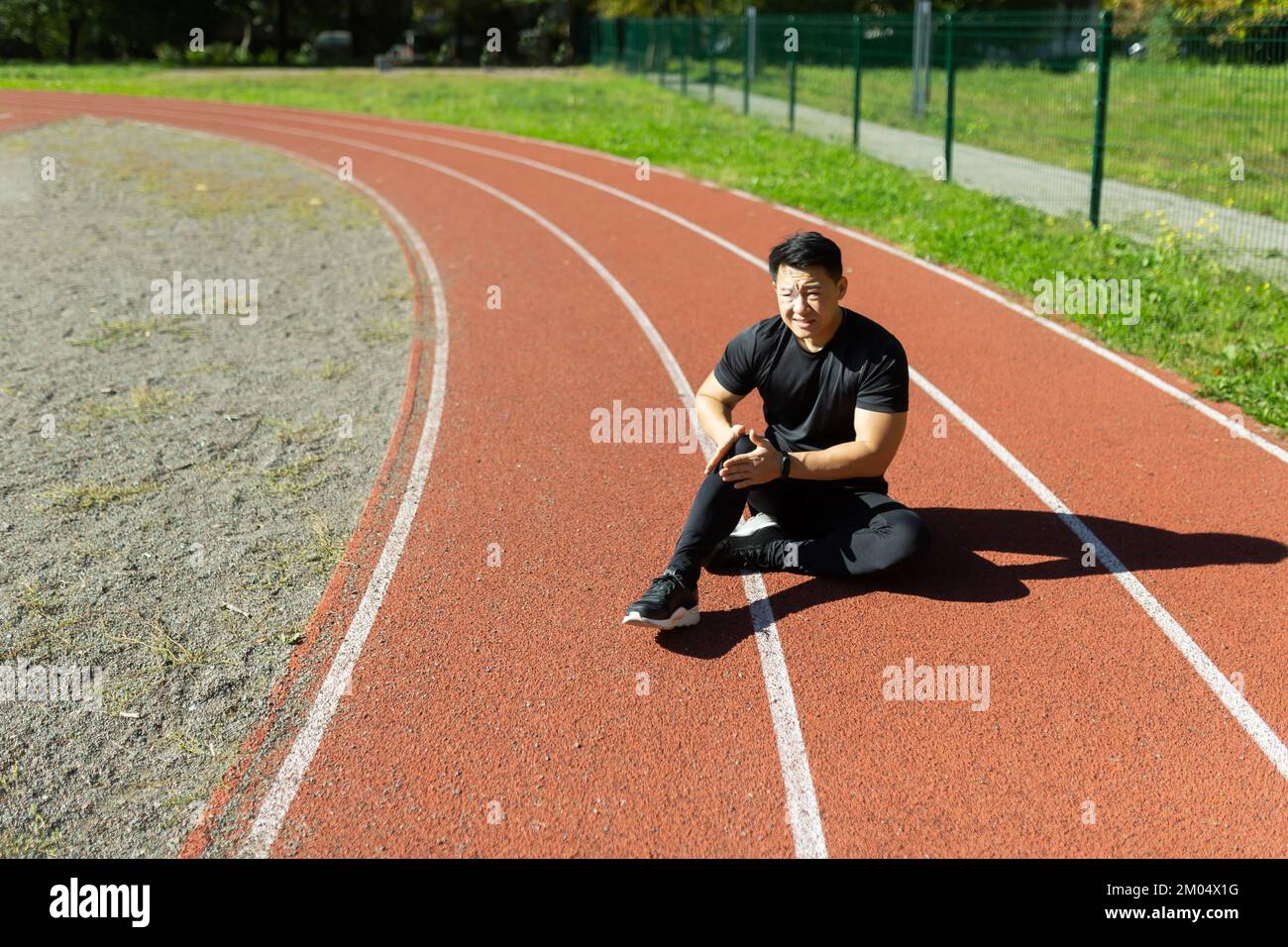 Sports injury. A young Asian man is sitting at the stadium on a treadmill, holding his leg and