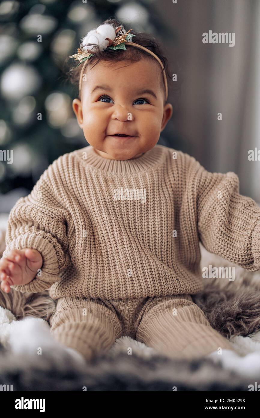 Happy mixed race baby girl sits on bed against background of Christmas tree. Concept of interracial family and unity between different human races. Stock Photo