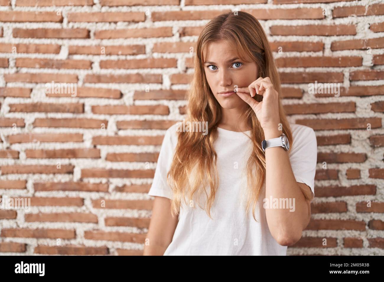 Young caucasian woman standing over bricks wall mouth and lips shut as ...