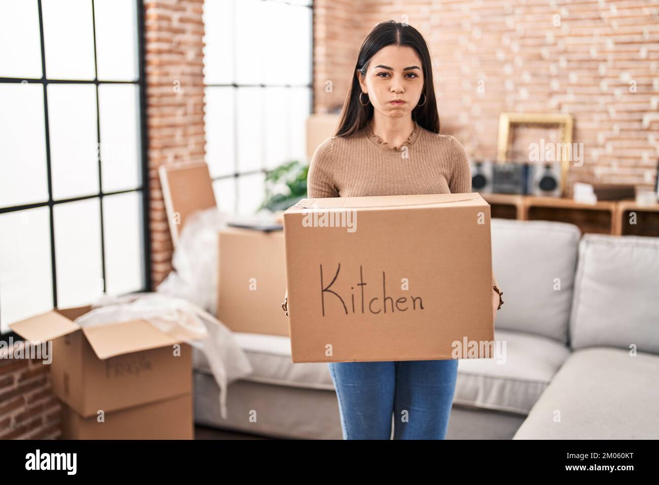 Young woman moving to a new home holding box puffing cheeks with funny ...