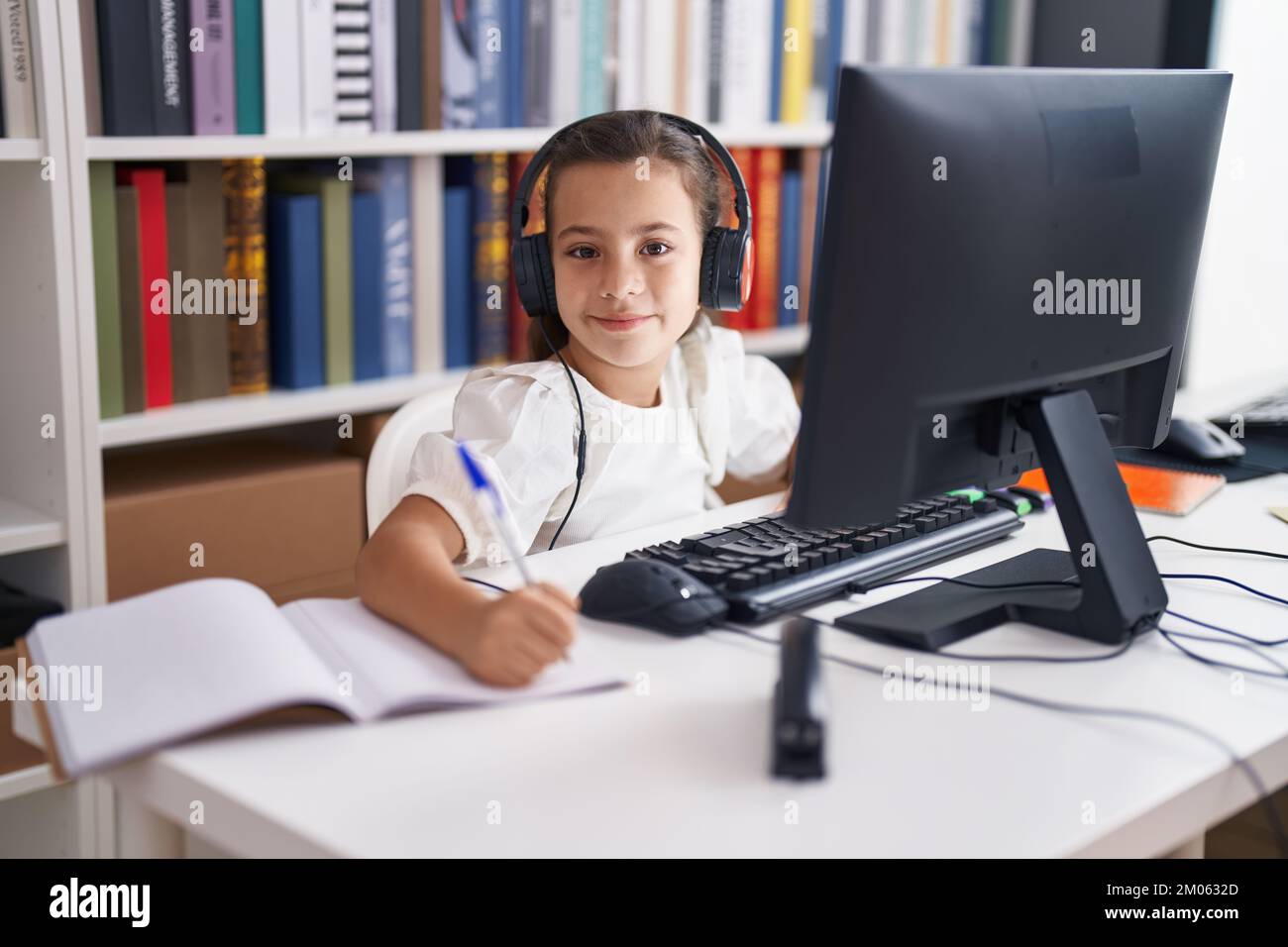 Adorable hispanic girl student using computer and headphones writing on ...