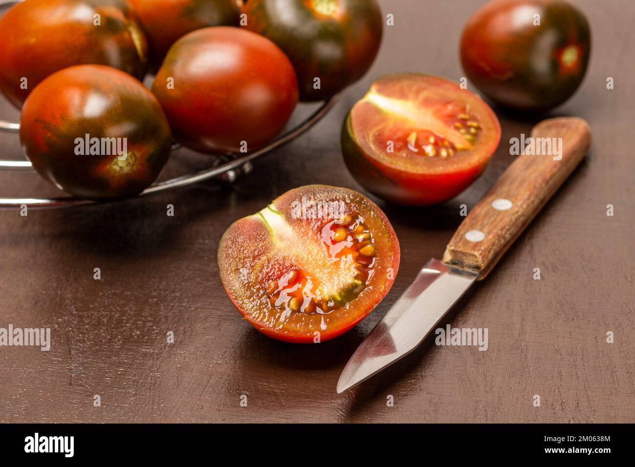 Knife and two halves of a tomato. Whole tomatoes on a metal stand. Top view. Brown background. Stock Photo