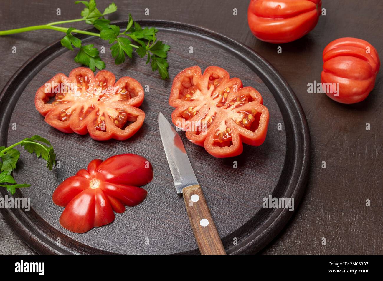 Sliced tomatoes and knife on cutting board. Two whole tomatoes on table. Top view. Brown background. Stock Photo