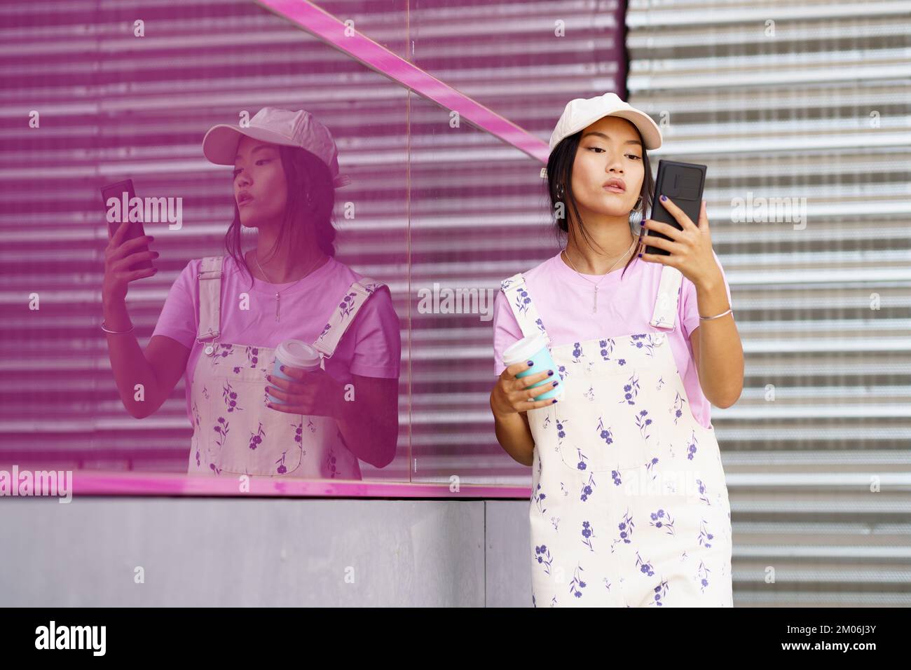 Young Asian woman with smartphone and coffee near pink wall Stock Photo