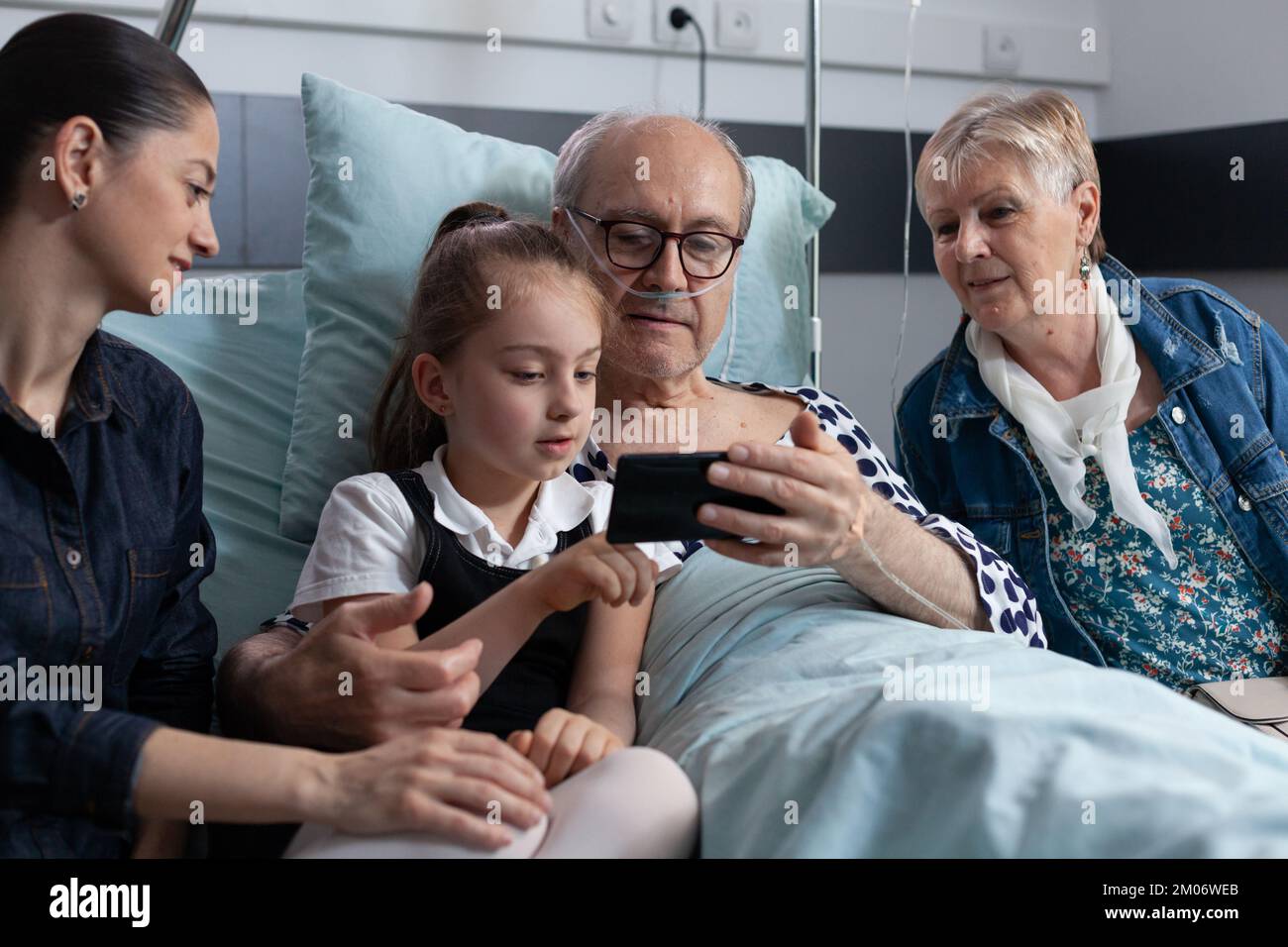 Little girl showing cell phone videos to grandfather hospitalized in geriatric clinic. Elderly man looking family photos at hospital room. Sick man relatives showing pictures on smartphone. Stock Photo