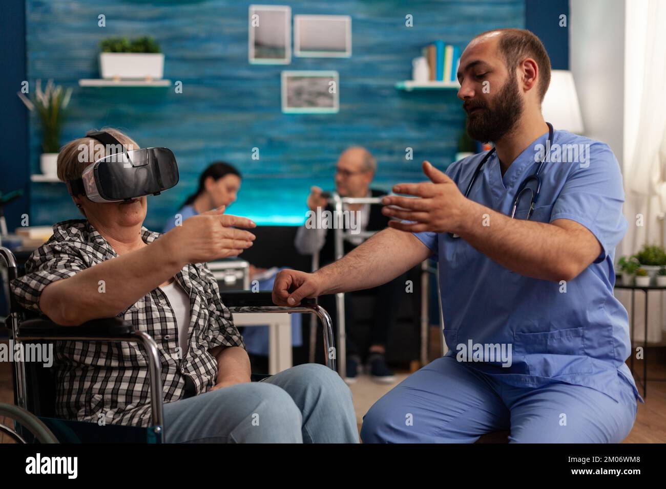 Elderly woman uses a virtual reality headset on a wheelchair in a nursing home to explore augmented reality. Male nurse in common room of skilled care facility helping patient to have some fun. Stock Photo
