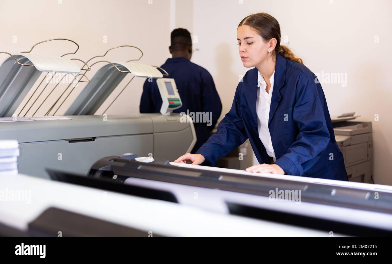 Concentrated young female typographer in a blue robe uniform pressing the buttons on the plotter in the typography Stock Photo