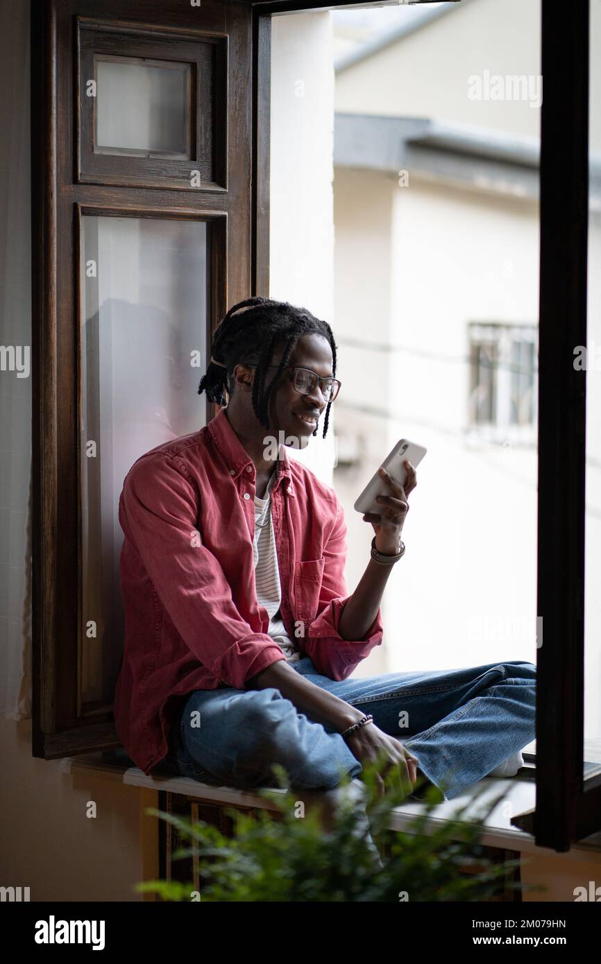 Happy carefree African American man sits on windowsill of apartment chatting with friends via phone Stock Photo