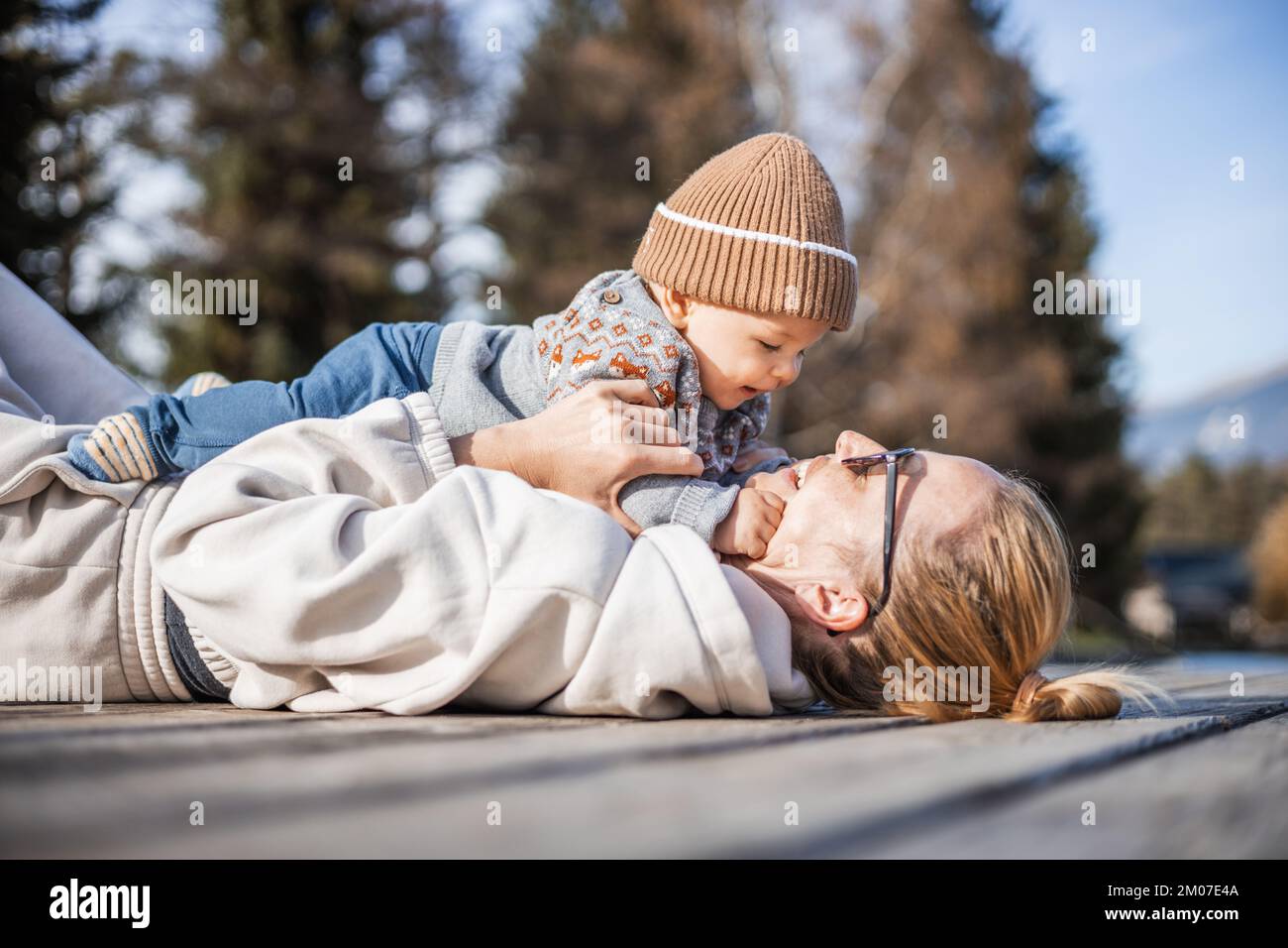 Happy family. Young mother playing with her baby boy infant oudoors on sunny autumn day. Portrait of mom and little son on wooden platform by lake. Positive human emotions, feelings, joy Stock Photo