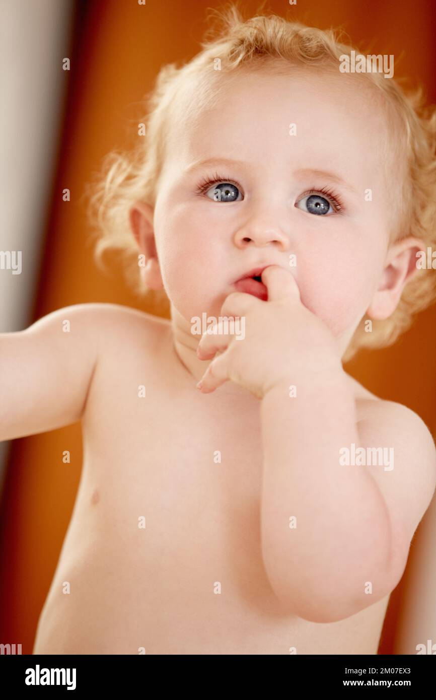Hmm, I wonder whats over there. Cute baby boy sucking on his finger while standing indoors. Stock Photo