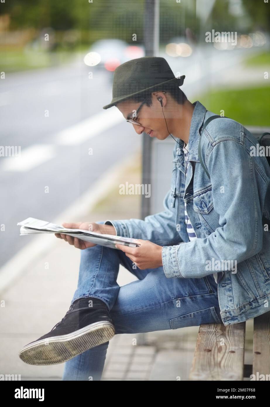 Reading todays newspaper. Hip young guy sitting at a bus stop while reading a newspaper. Stock Photo